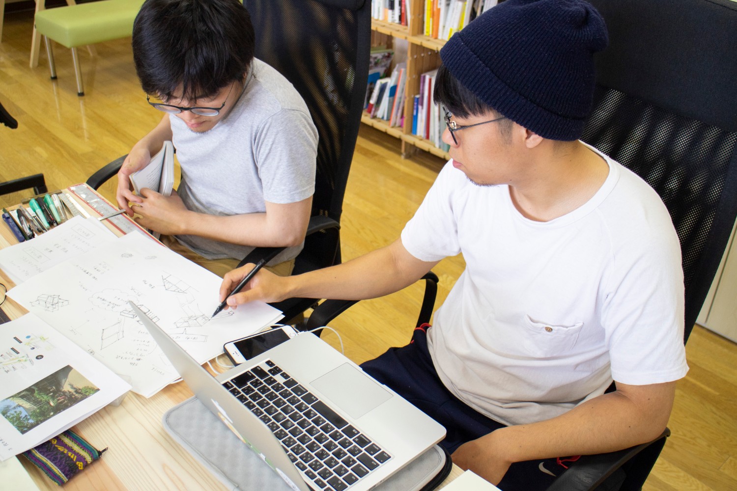 平山さんがノートパソコンの前でスケッチにペンを走らせ、隣の男性と作業をしている写真