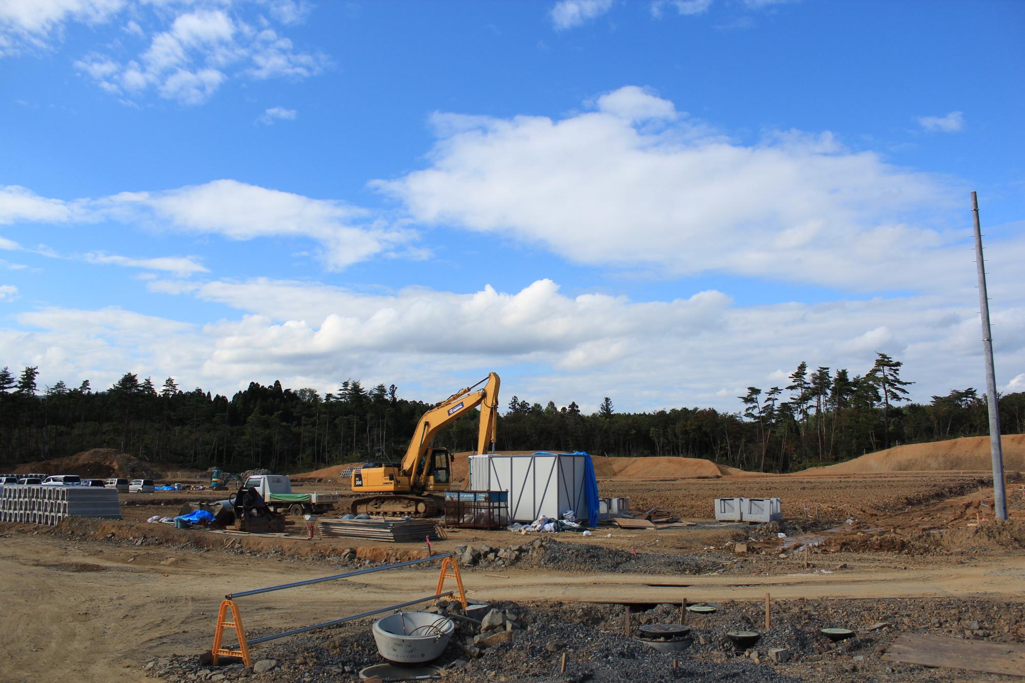 重機と建設資材が点在する空き地で、青空の下で掘削作業が行われている造成工事中の写真