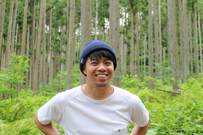 白いTシャツと青いニット帽をかぶった平山さんが深い森林の中で笑顔を見せて立っている写真