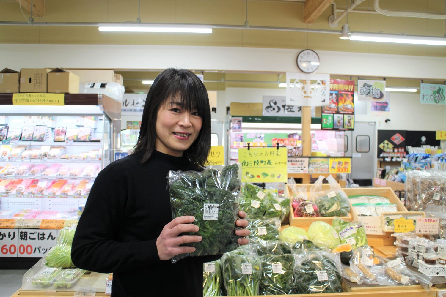 新鮮な野菜が並んでいる陳列棚の前で、笑顔で透明袋に入った野菜を両手で持つ中村店長の写真