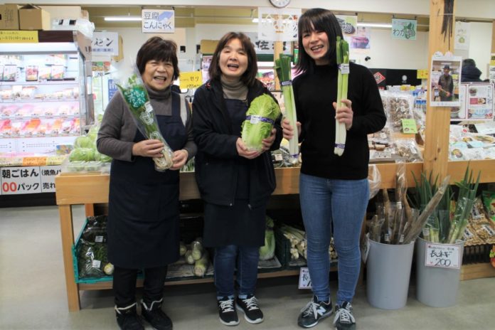 三人の女性が店内で笑顔で並び、それぞれ長ねぎや白菜などの新鮮な野菜を手に持っている様子の写真