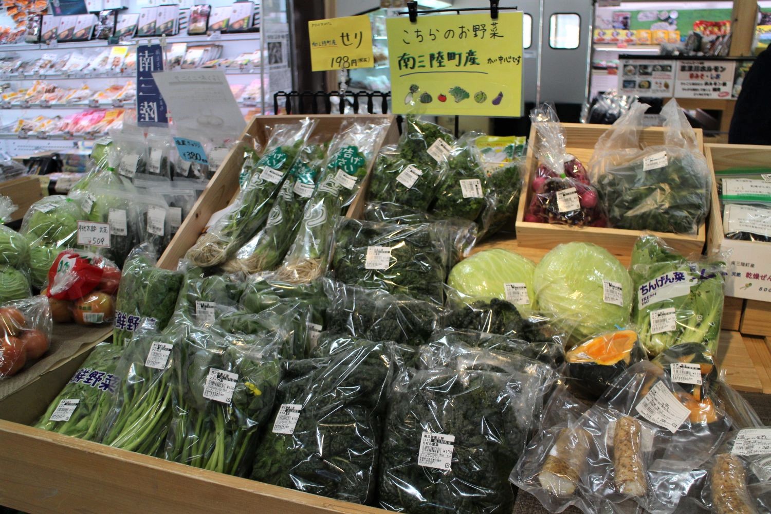 地元・南三陸町産の新鮮な野菜が袋詰めで並ぶ陳列台で、キャベツやセリなど多様な野菜が売られている写真