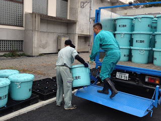 作業着を着た二人の男性が、屋外で青い容器をトラックに積み込んでいる様子の写真
