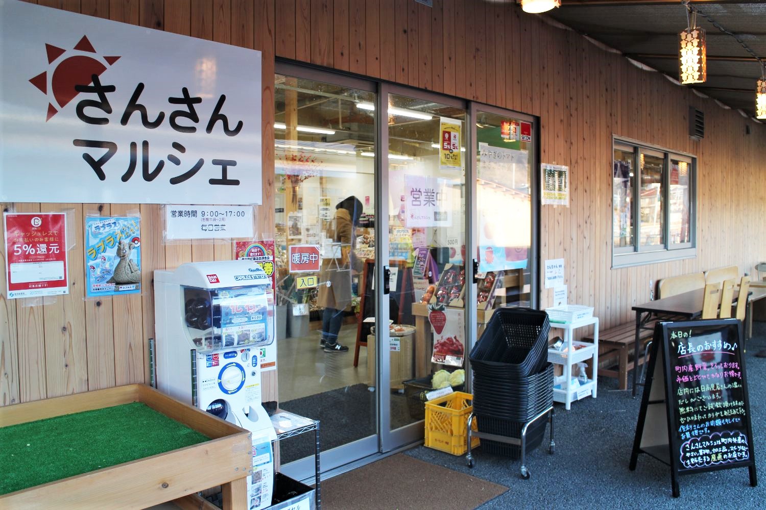 木目の外壁に「さんさんマルシェ」と書かれた看板が掲げられた店舗の入口で、店内の様子や買い物かごなども見える写真
