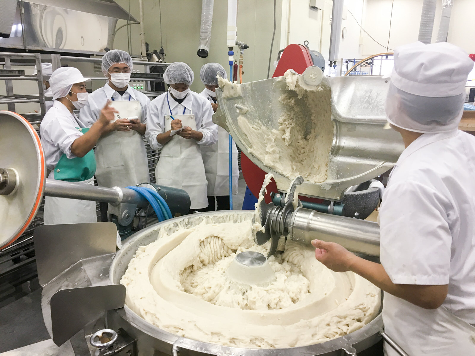 白衣と帽子をかぶった学生たちが白いすり身が入った大きな機械の前で職員の説明を受けながらメモを取っている実習の様子の写真