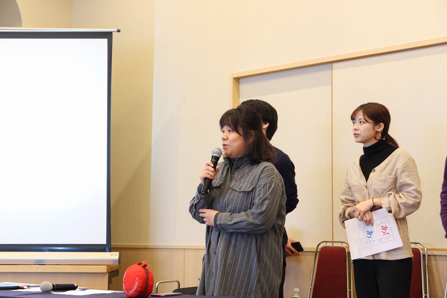 スクリーンの横でマイクを持って話している城戸裕子先生、その後ろに3人の関係者が立っている写真