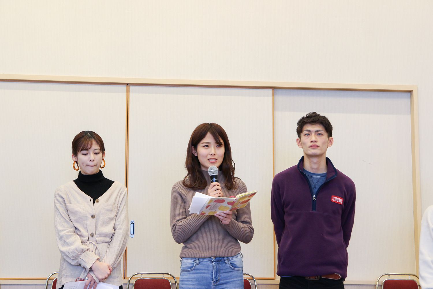 服部星来さんがマイクを持って紙を読んでおり、左側に女性、右側に男性が立っている写真