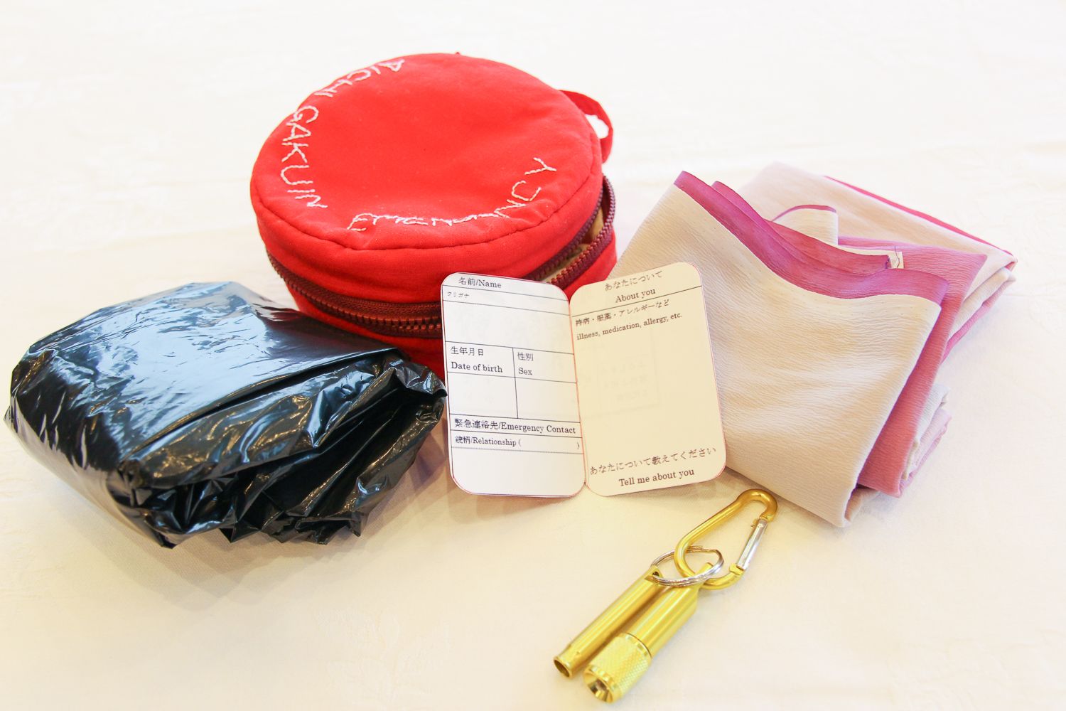 赤い防災ポーチ、黒いビニール袋、個人情報カード、金色のホイッスルとピンク色のふろしきの写真