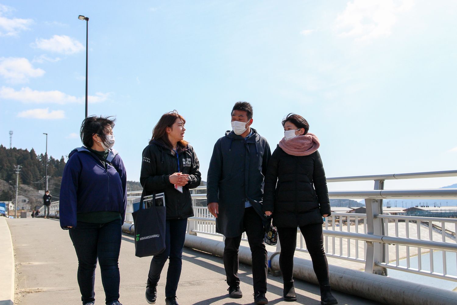 青空と遠くの山が見える橋の上を、3人の男女が左から2番目の女性の話を歩きながら聞いている写真