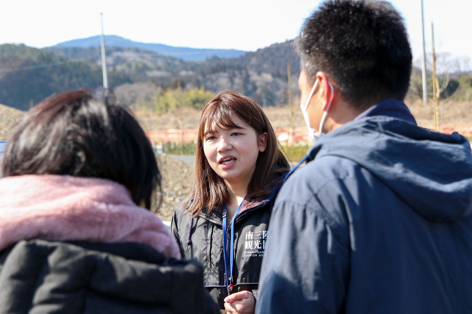 山に囲まれた屋外で、黒いジャンパーを着た女性の話しを、参加者の人達が耳を傾けている写真