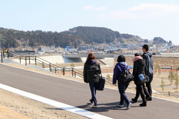 青空の下、遠くには住宅や山が見えている公園のなだらかな遊歩道を、四人の男女が歩いている後ろ姿の写真
