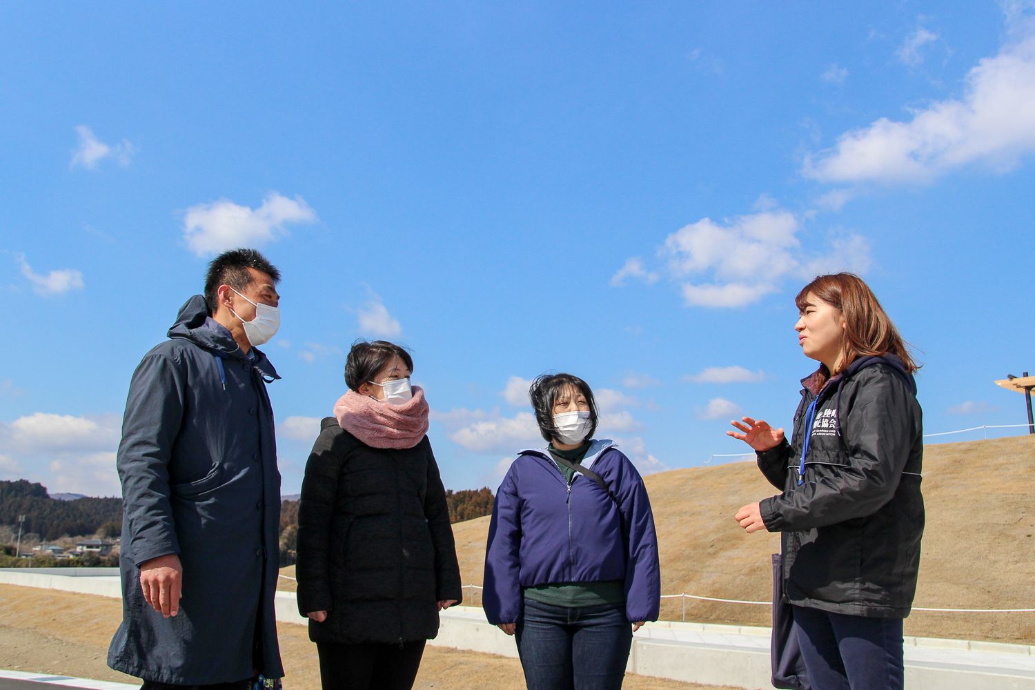 青空の下、黒い観光協会のスタッフジャンパーを着た女性の話を、向かい合って立っている参加者の3人が聞いている写真