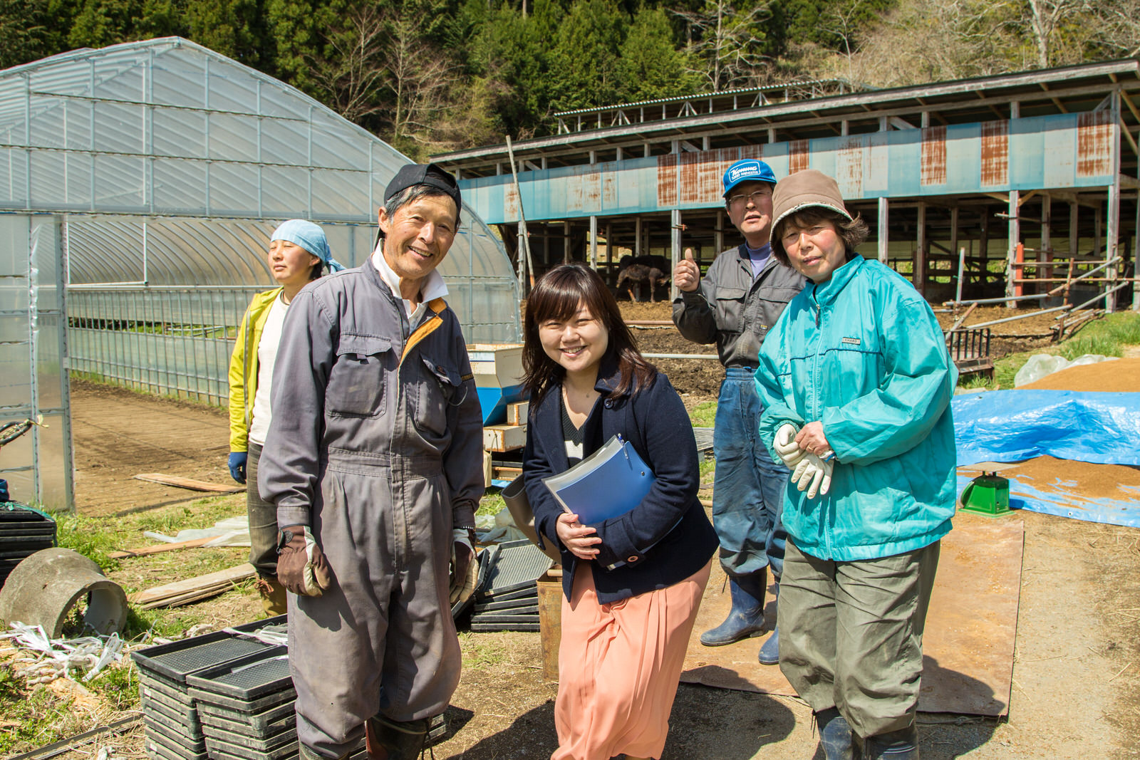 リポーターの女性と阿部勝善さん、他男性2人と女性1人がビニールハウスと牛小屋を背景に立っている写真
