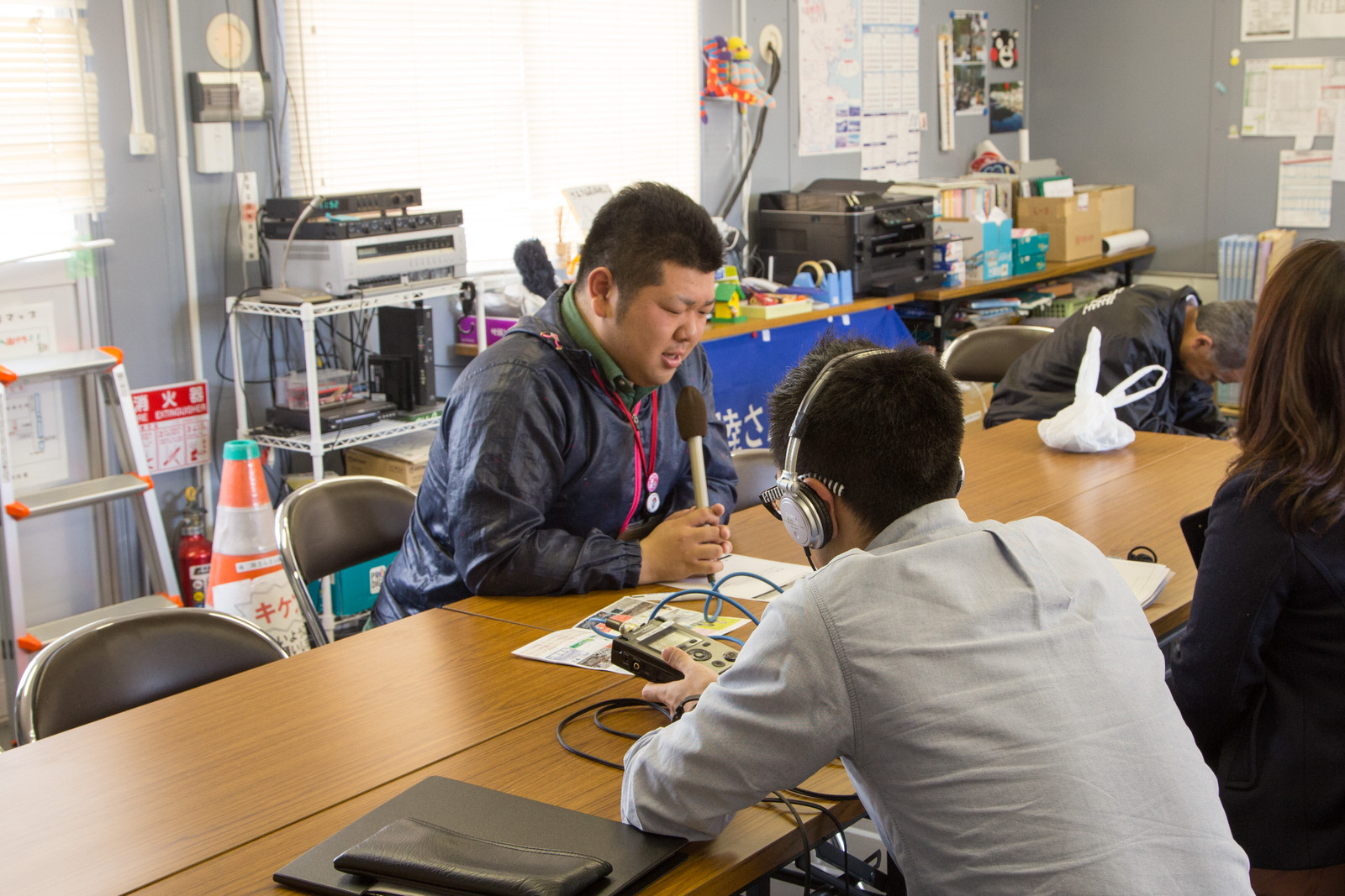様々な機材などが置かれた室内で、ヘッドホンを着けて機械を操作している男性とリポーターの女性と向かい合って、長机の席に座った佐藤さんが緊張した面持ちでマイクを持って話をしている様子の写真