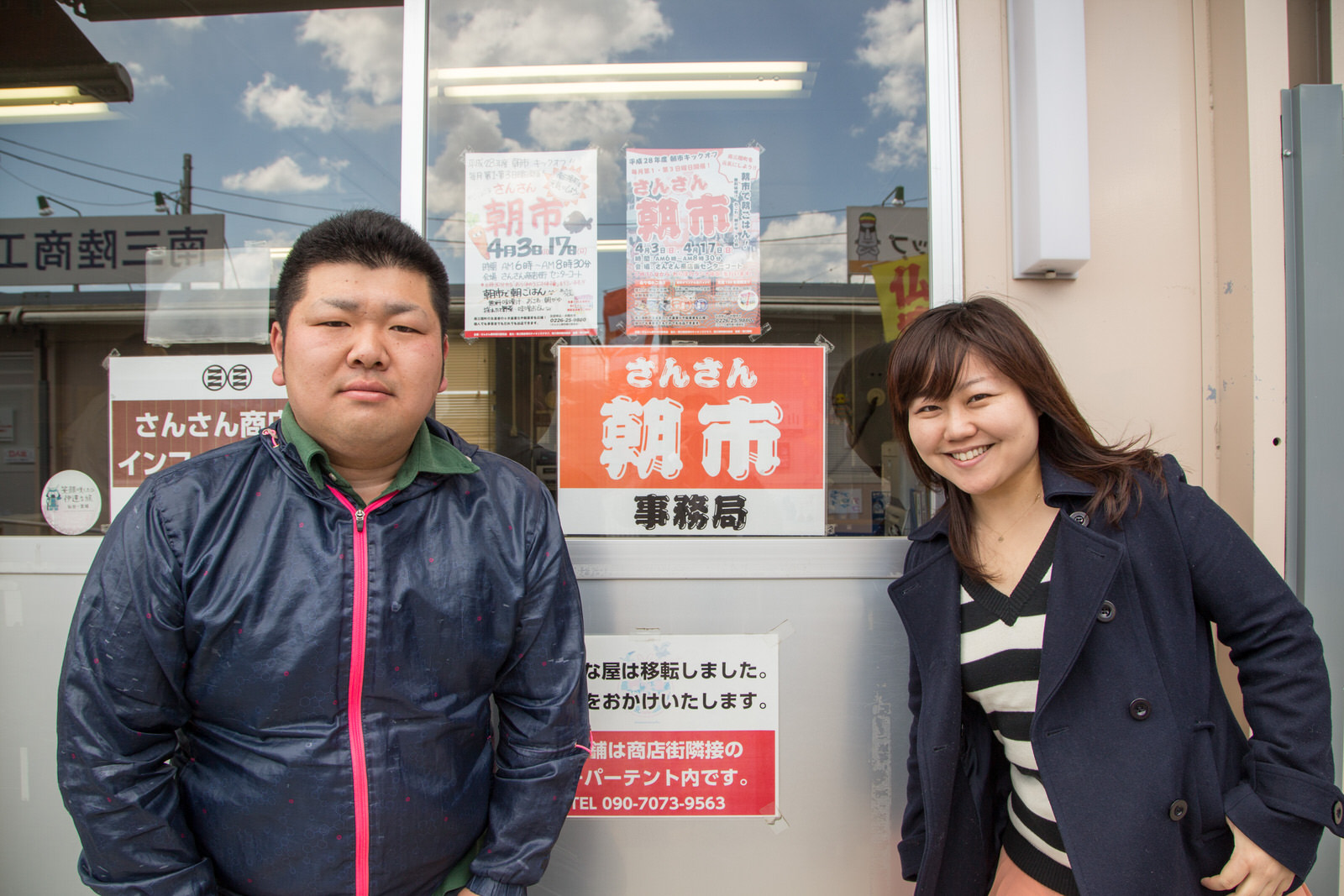 「さんさん朝市事務局」など複数の張り紙が貼られた入り口前に立つ、南三陸町商工会の佐藤潤也さんとリポーターの女性の写真