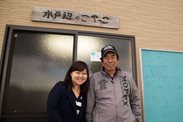 「水戸辺のこやっこ」と手書きで書かれた木の看板が取り付けられた建物の入り口前で、リポーターの女性と戸倉漁師の会松岡孝一さんが笑顔で並んで立っている写真