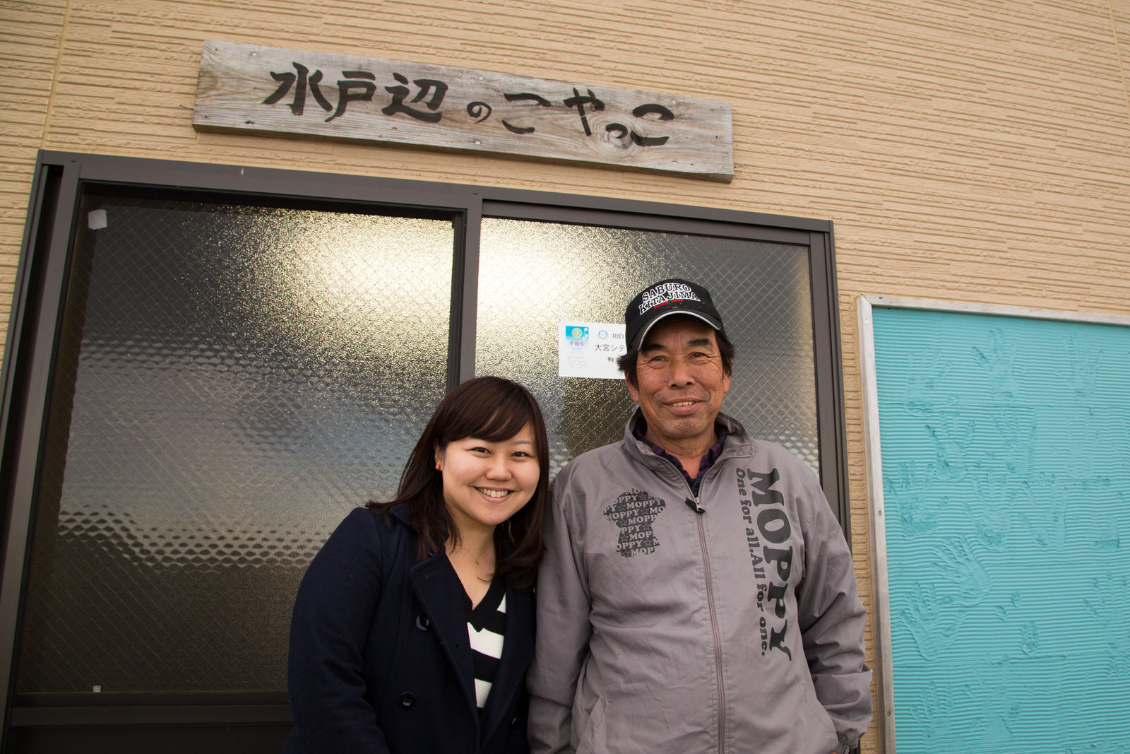 「水戸辺のこやっこ」と手書きで書かれた木の看板が取り付けられた建物の入り口前で、リポーターの女性と戸倉漁師の会松岡孝一さんが笑顔で並んで立っている写真