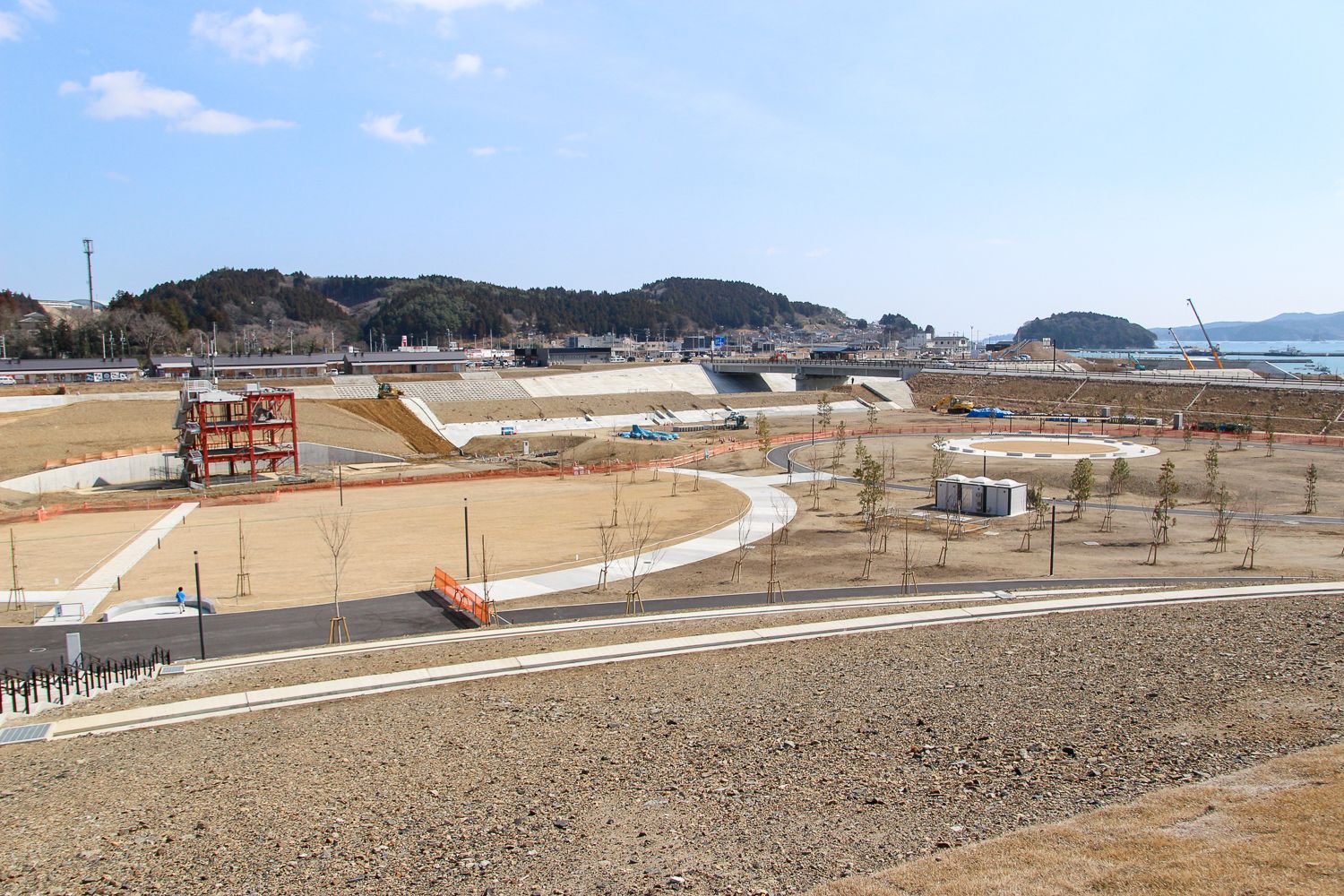 遠くには町と海が見える、造成中の広い復興祈念公園に遊歩道や間隔をあけて植えられた若木が立ち並んでいる全景の写真