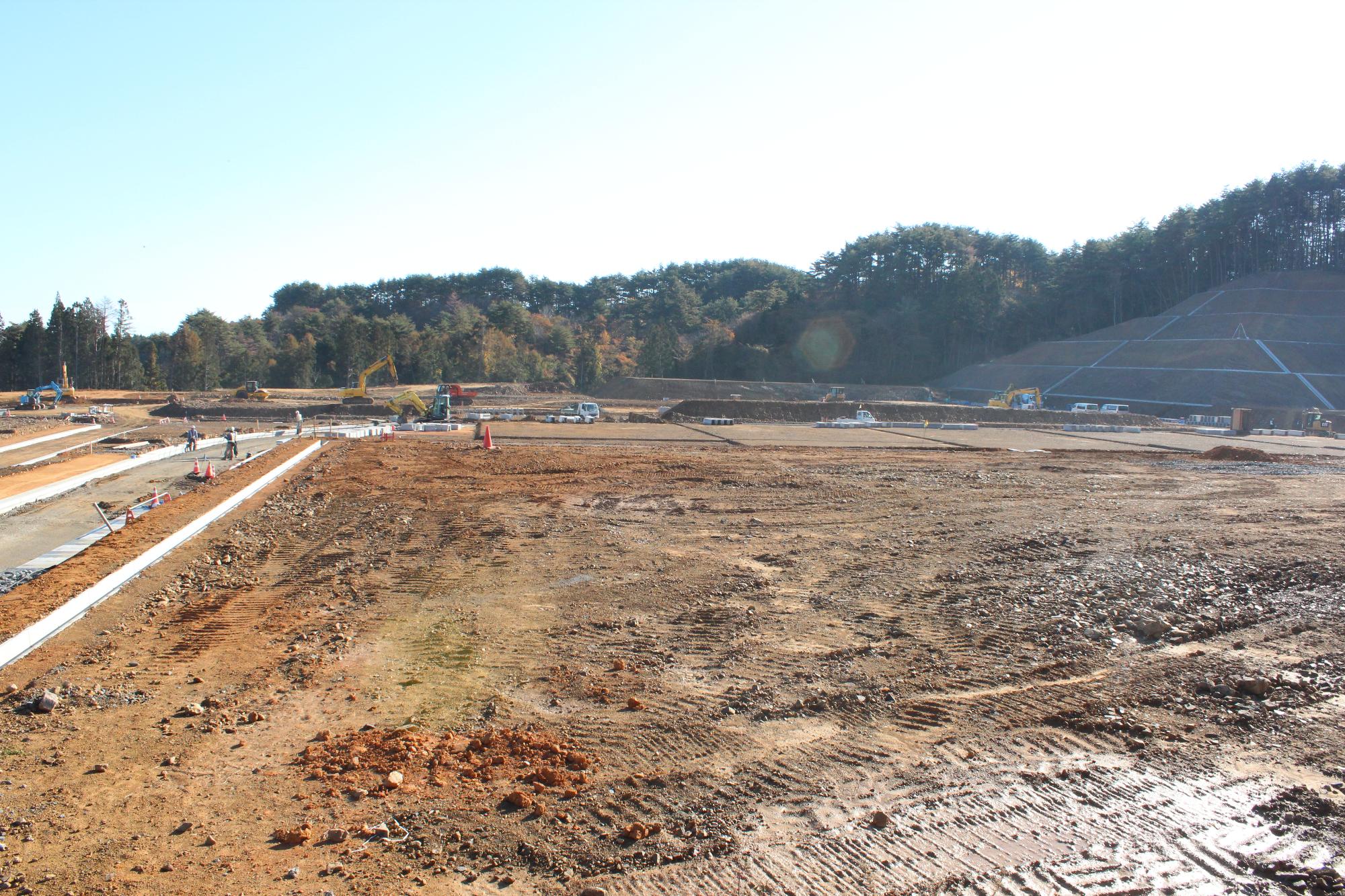 背景に森林と法面工事の様子が見え、舗装準備が進み、重機や作業員が整然と配置されている造成地の写真