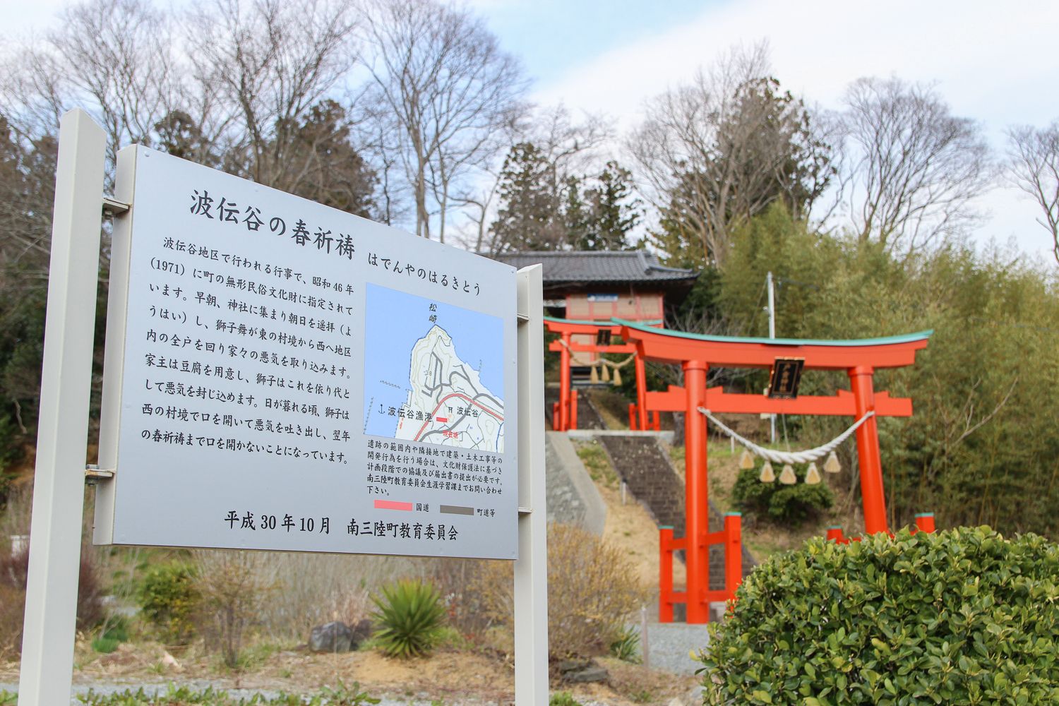 「波伝谷の春祈祷」と書かれ行事の内容や地図が説明されている案内板が、朱色の2基の鳥居と神社の本殿の前に立っている様子の写真