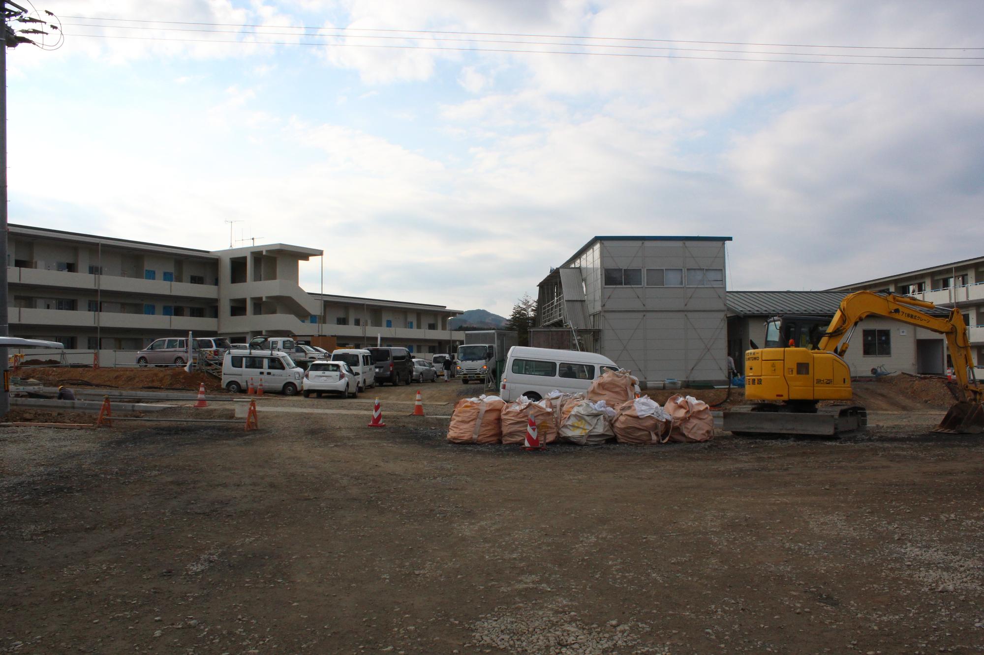 仮設建物の横で黄色いショベルカーが動き、人々が作業する様子が見える建設現場の写真
