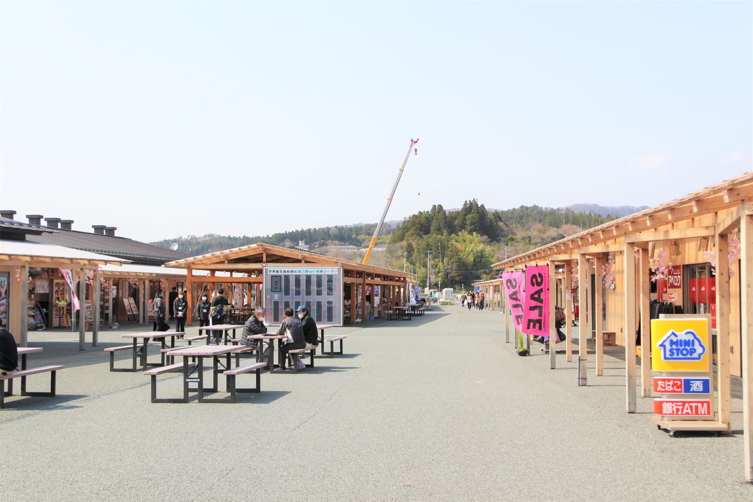 背景には山や木々が広がっており、広場にある木造の商店街と自動販売機があるさんさん商店街の写真