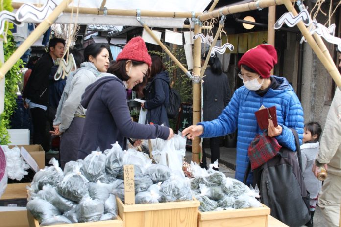 古民家が立ち並ぶ細い通路に設置された販売ブースで、赤いニット帽を被った女性がビニール袋に入ったワカメを購入している様子の写真