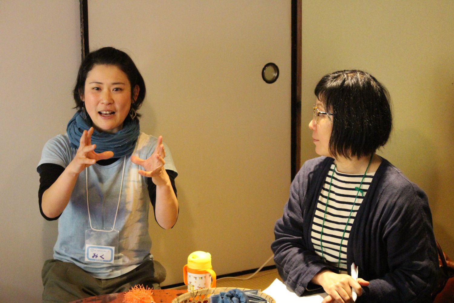 室内の襖の前で、青いスカーフを巻いた女性が身振り手振りで話し、隣でボーダーシャツの女性がメモを取りながら聞いているお話会の様子の写真