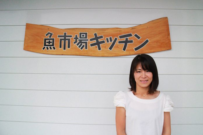 「魚市場キッチン」と黒い文字で書かれている木の看板が設置された白い壁の前に白い服を着た中村悦子さんが笑顔で立っている写真
