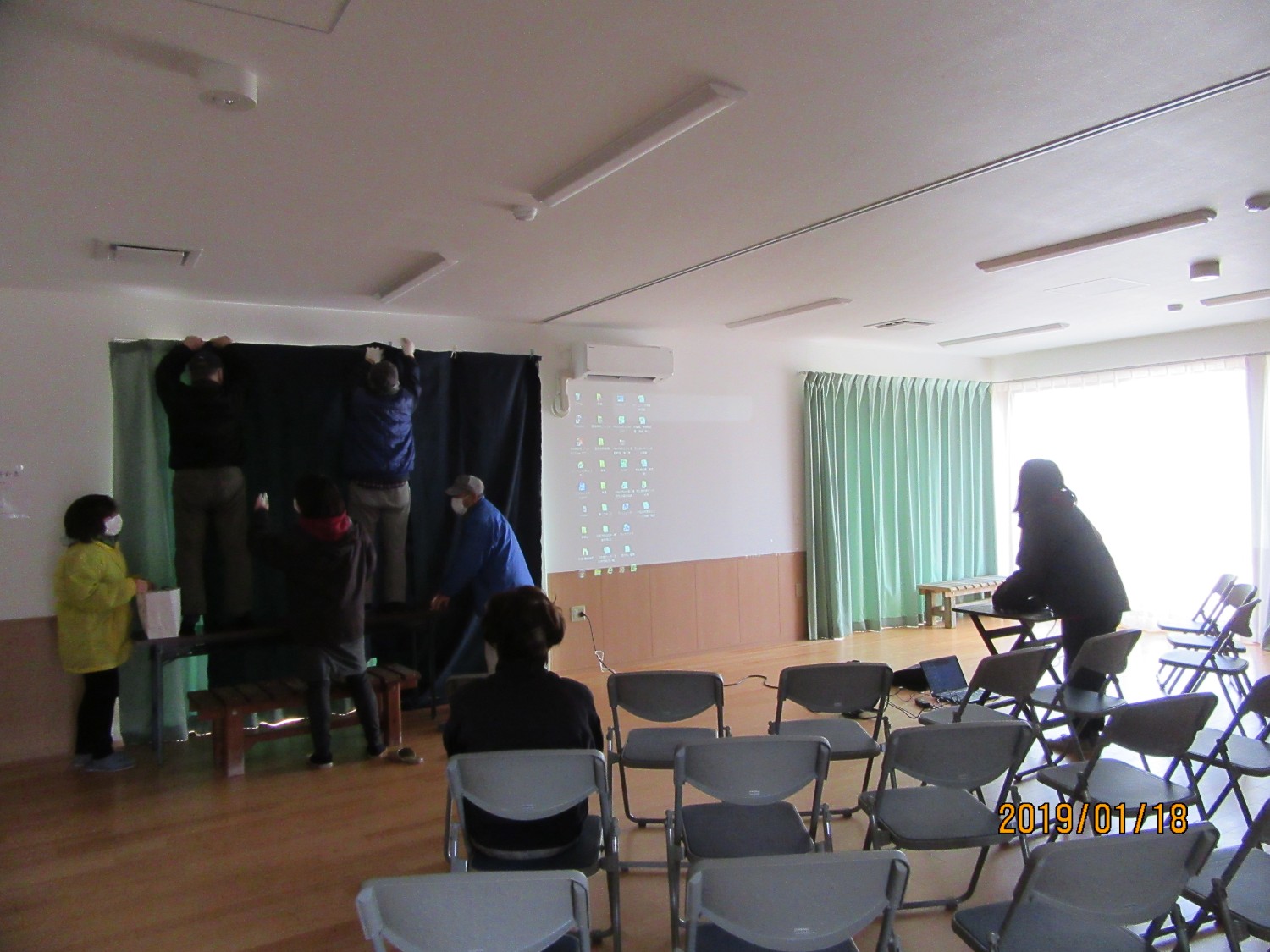 数人が部屋の前方でカーテンを取り付け、手前には椅子が整然と並べられ、右側にはプロジェクターの画面が壁に映し出されている写真