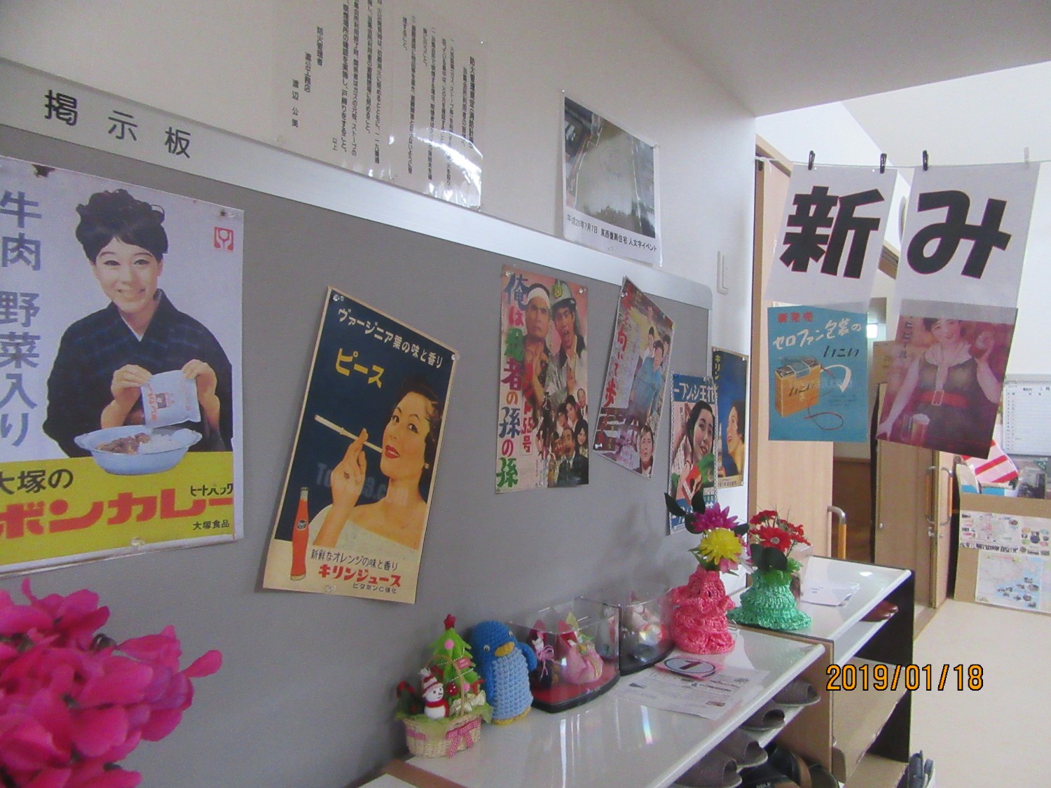 掲示板に数枚のポスターが貼られ、手前の机の上には花や小物が丁寧に配置されている写真