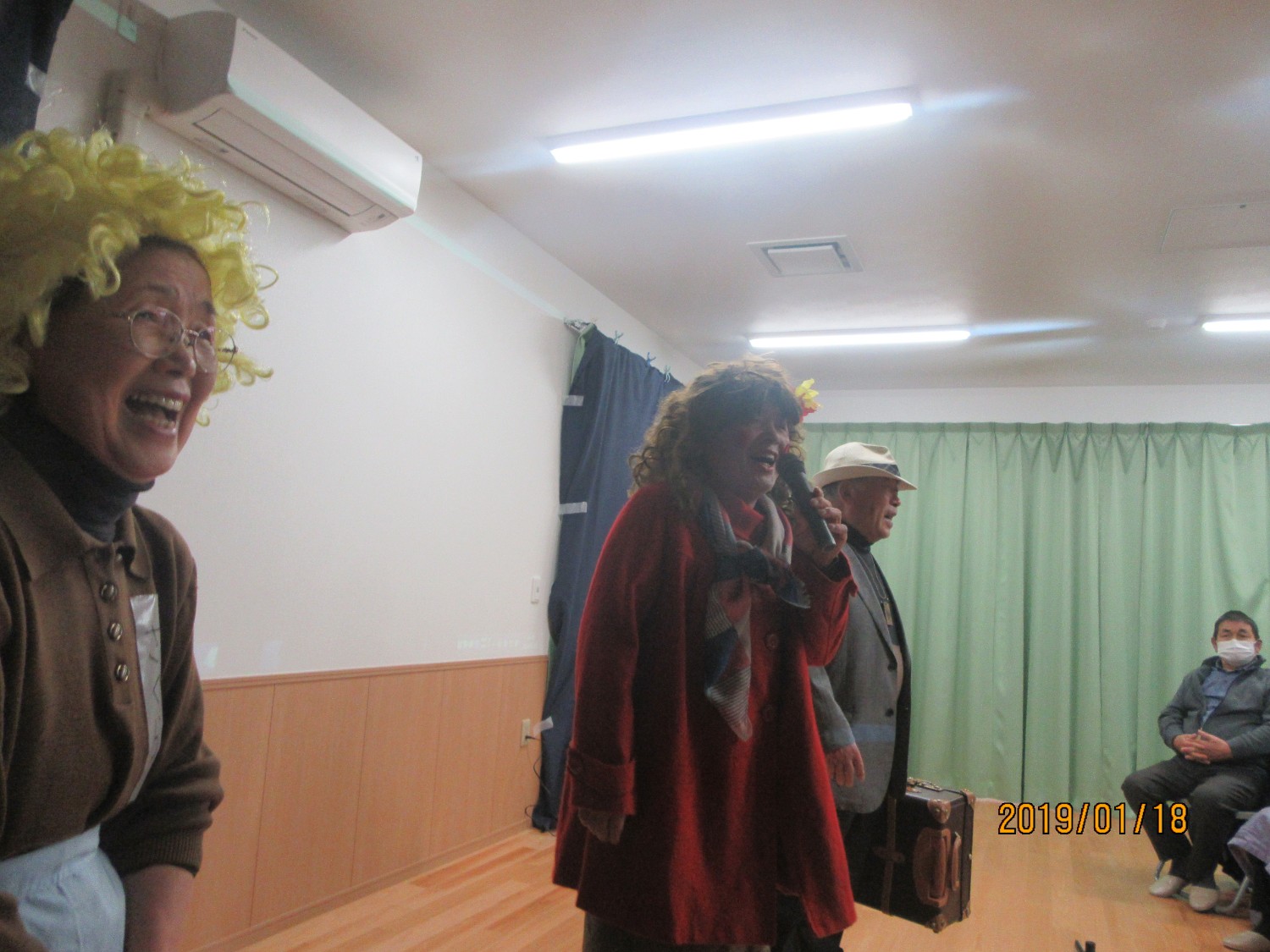 舞台の上で複数の人々がカツラをかぶって衣装を身にまとい、何らかの演劇を演じている様子が写っている写真