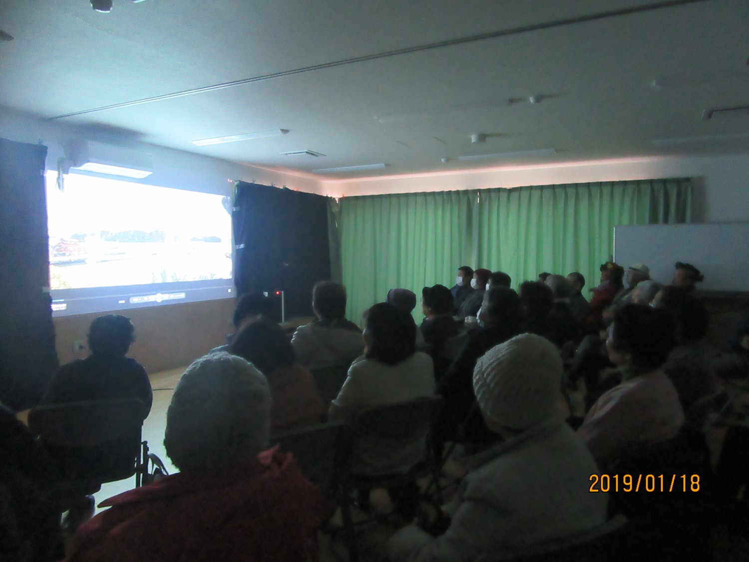 カーテンが閉められ暗くなった部屋の中で多数の人々が椅子に座って前方のスクリーンに映し出された映像を静かに見ている写真