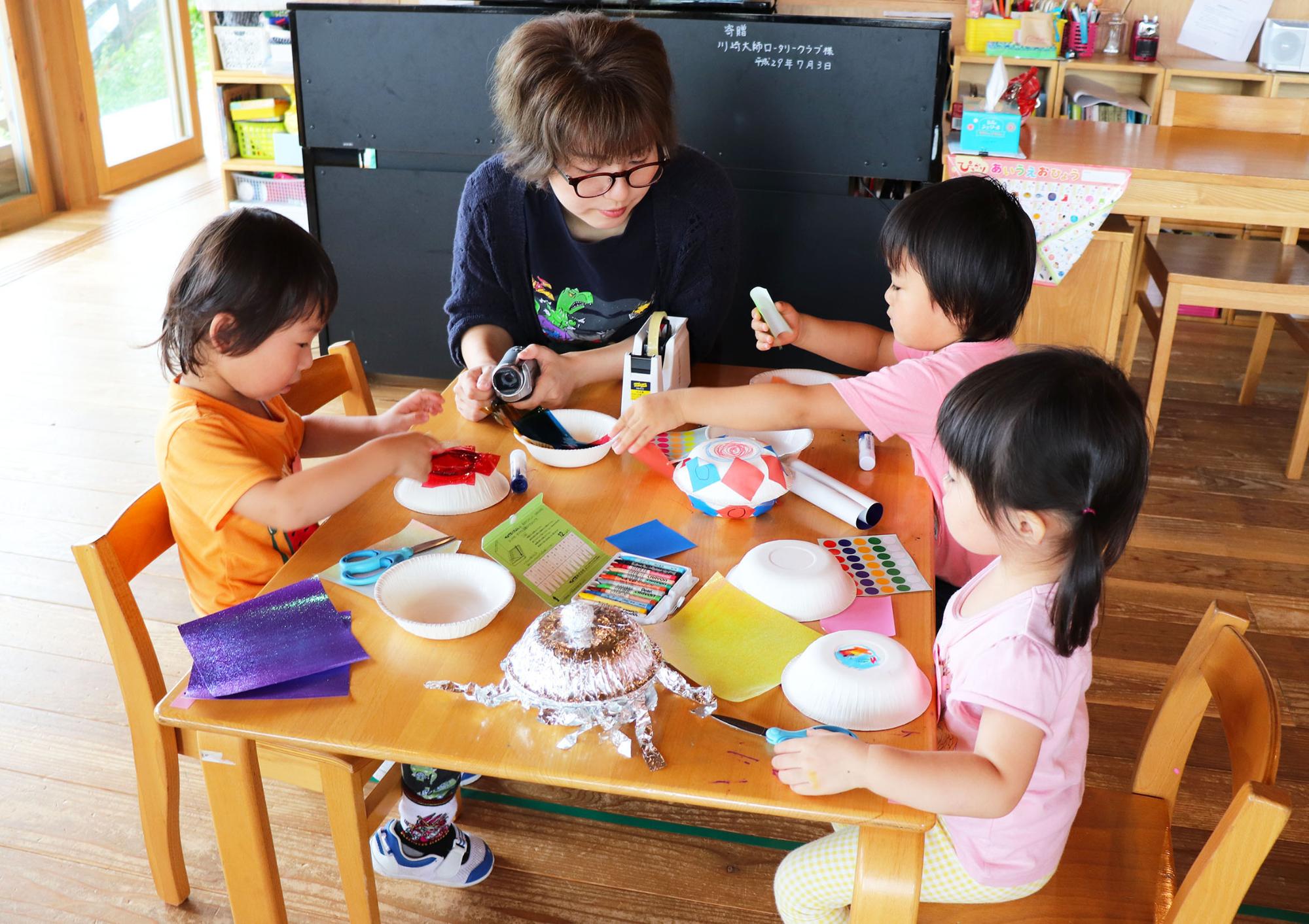 メガネをした先生が見守る中、小さな子供3人が椅子に座り工作を楽しんでる写真