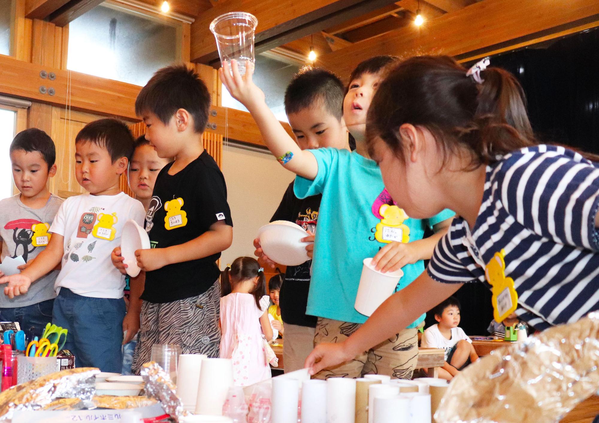 室内のテーブルの周りにたくさんの子どもたちが集まり、プラスチックコップや紙コップを手に取ったりして材料を選んでいる様子の写真