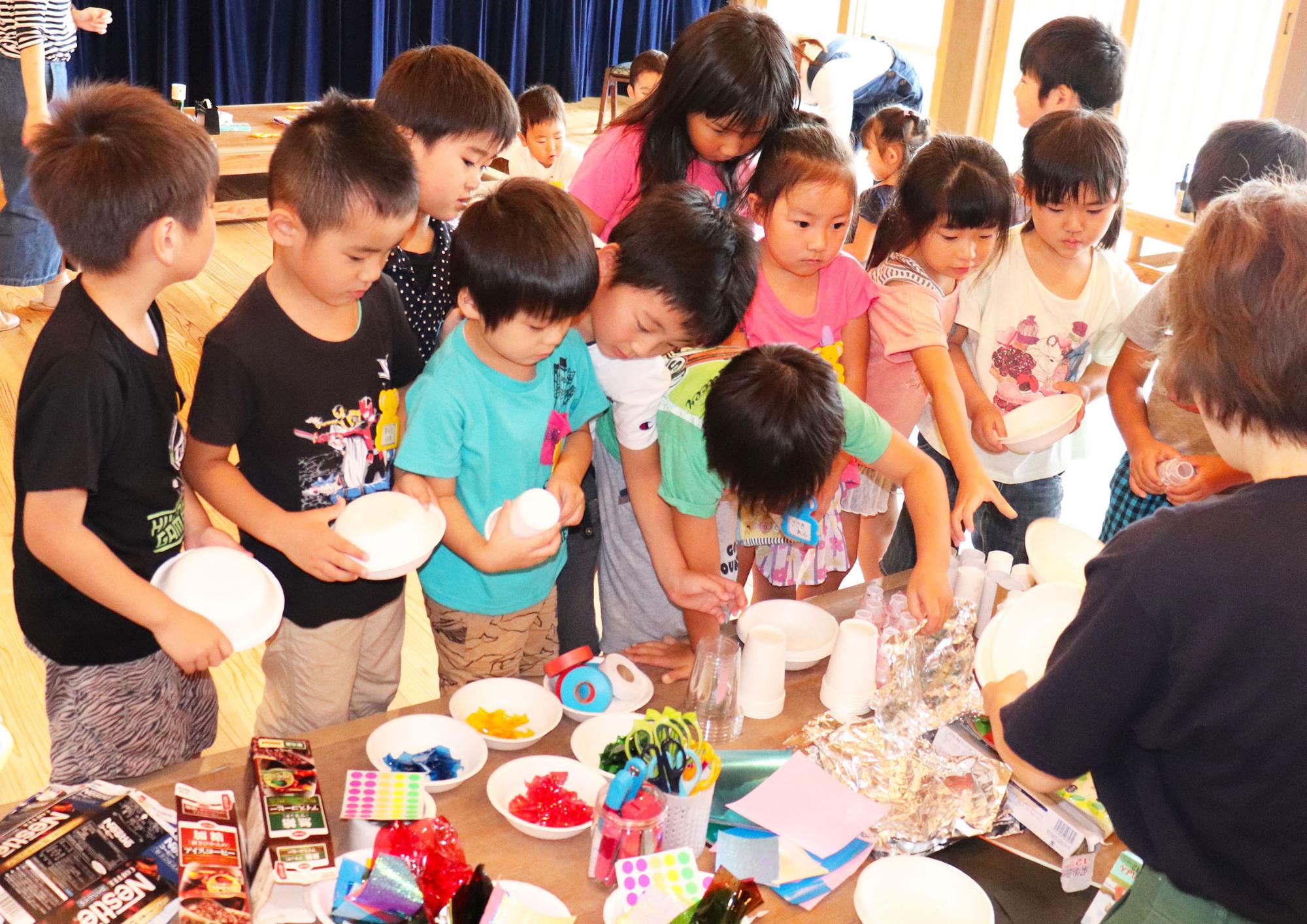 室内のテーブルの周りにたくさんの子どもたちが集まり、テーブルに置かれた材料を選んでいる様子の写真