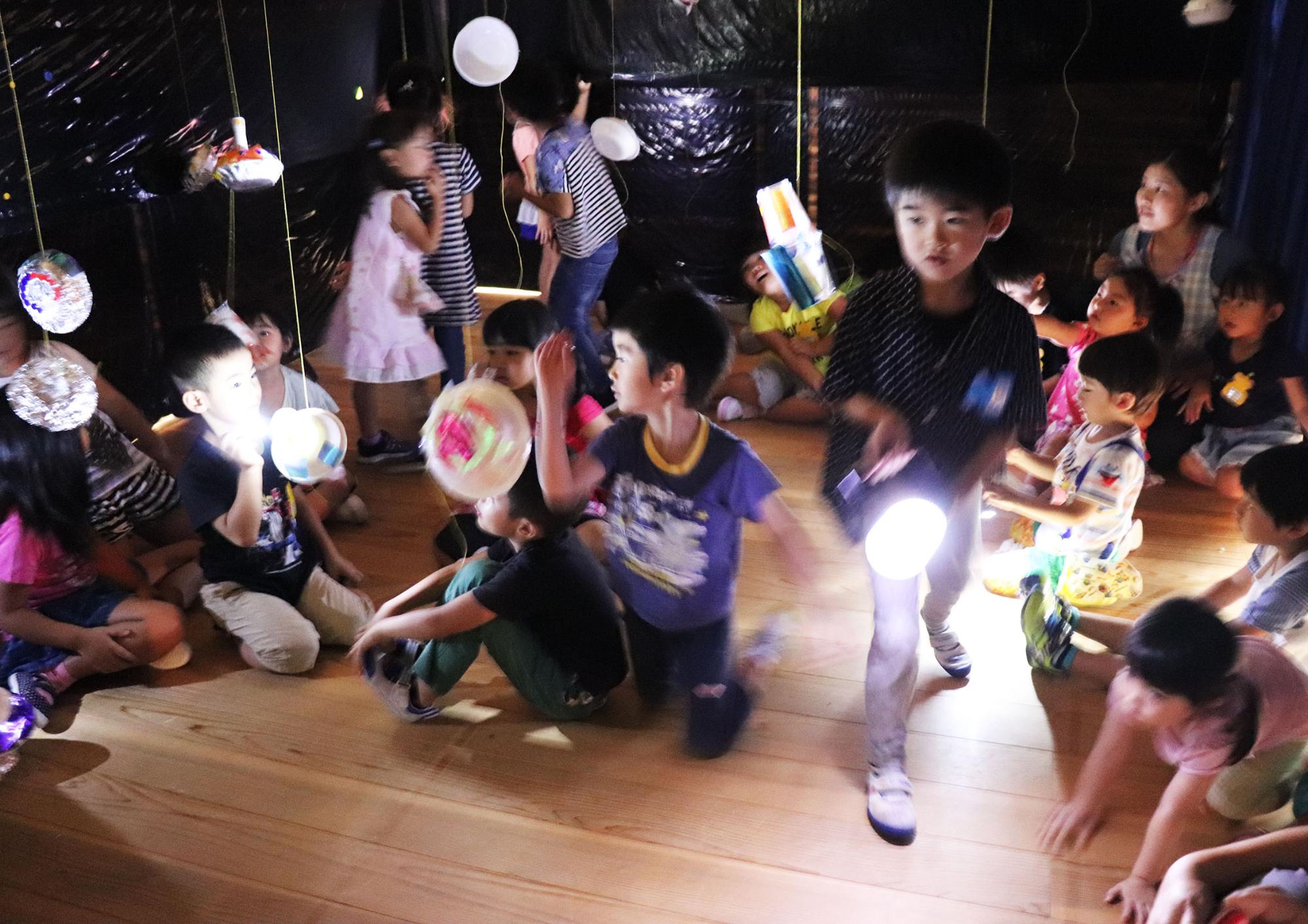 部屋の中が暗くされ、天井から糸で吊るした作品が吊るされ、たくさんの子どもたちが懐中電灯を手に楽しそうな様子の写真