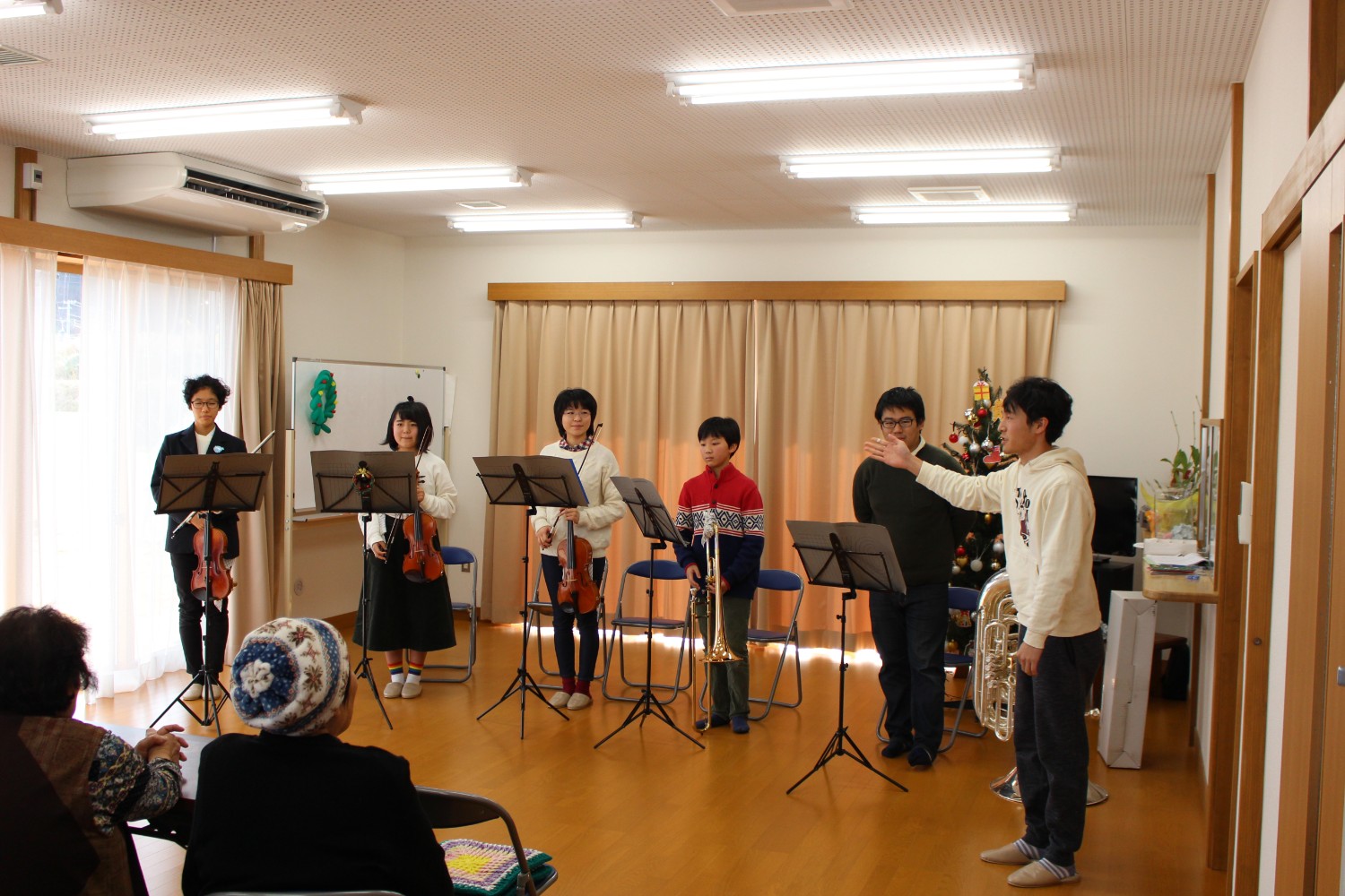 ヴァイオリンやトロンボーン、チューバなどをメンバー5人が譜面台の前に並び、右側では男性が手を広げて説明している様子の写真