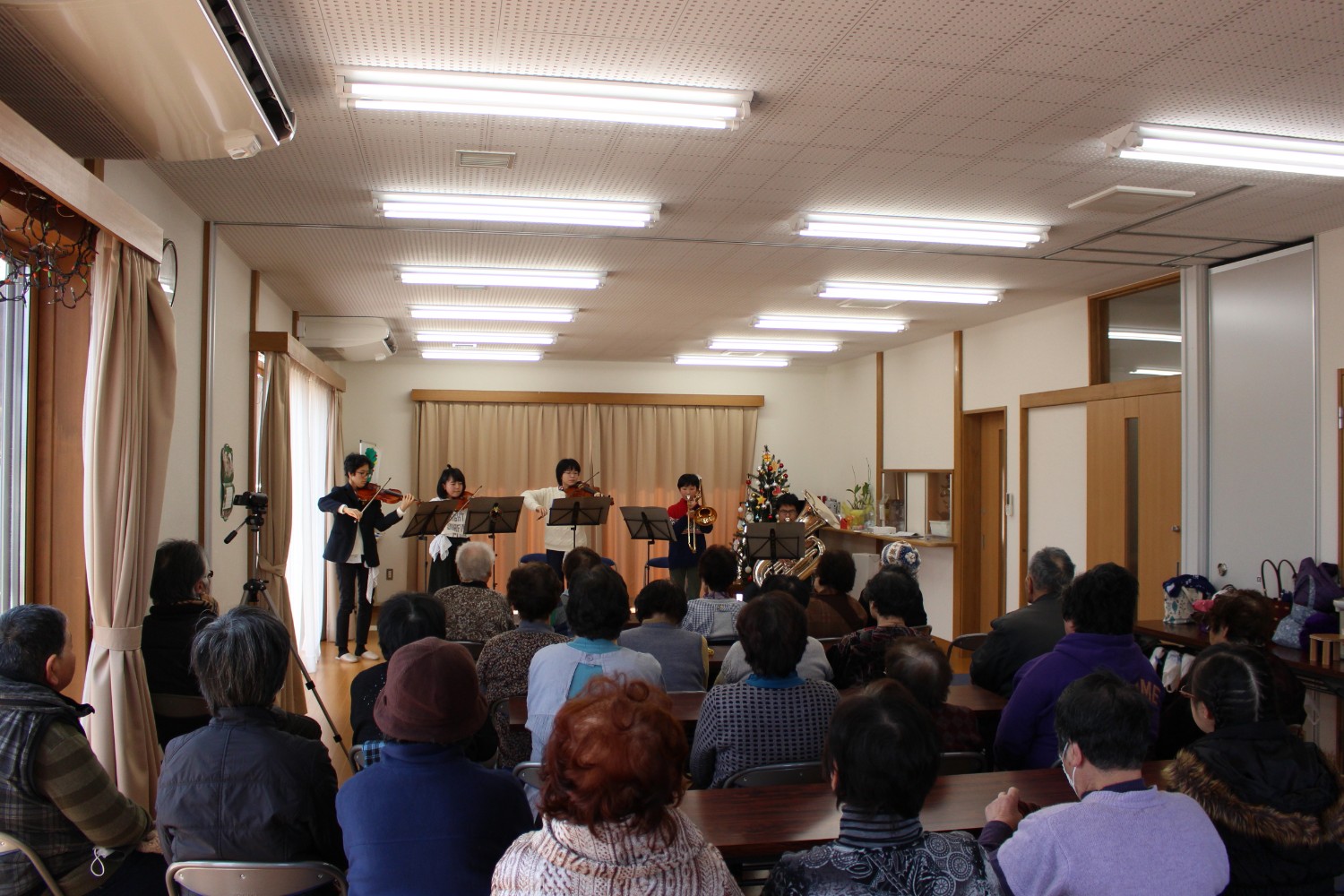 多くの高齢者が椅子に座って聴いている前で、東北ユースオーケストラのみなさんが弦楽器や金管楽器を使って演奏している様子を後方から写した写真