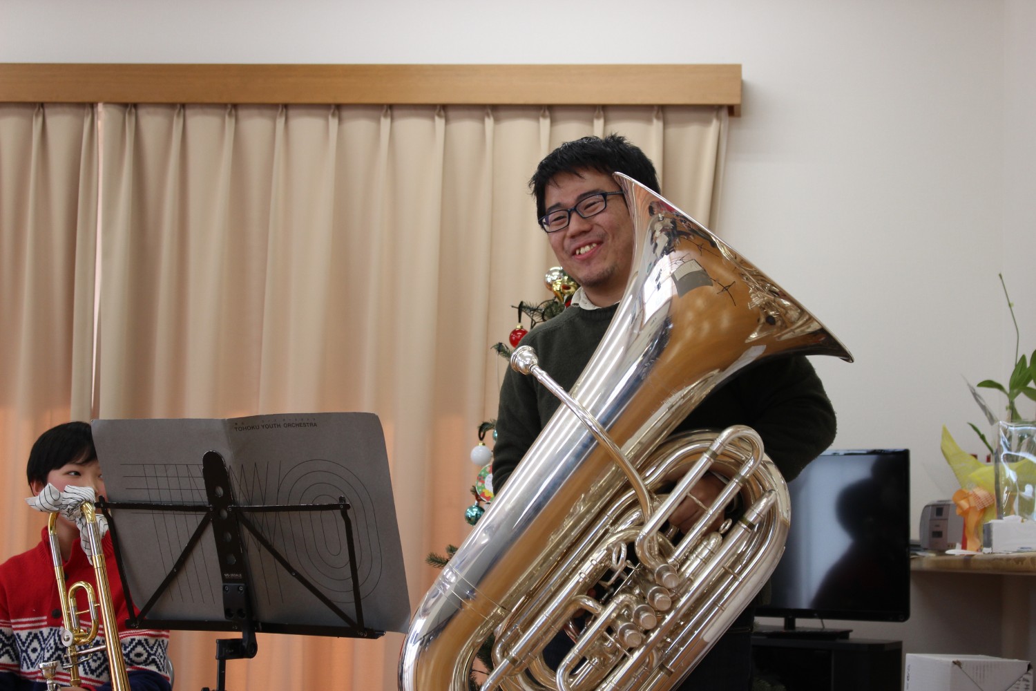 クリスマスツリーの前で、大きなチューバを抱えた冨澤悠太さんがにこやかに笑っている写真