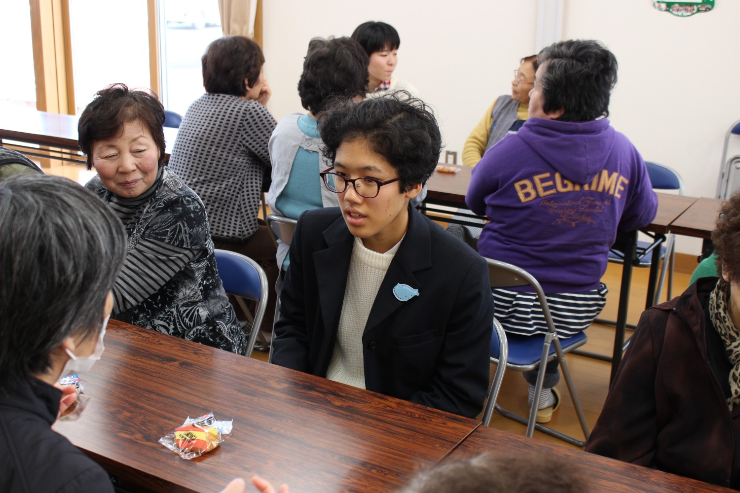 テーブル席で、眼鏡をかけたメンバーが笑顔で高齢者と向かい合って話しており、まわりでもメンバーと高齢者が交流している様子の写真