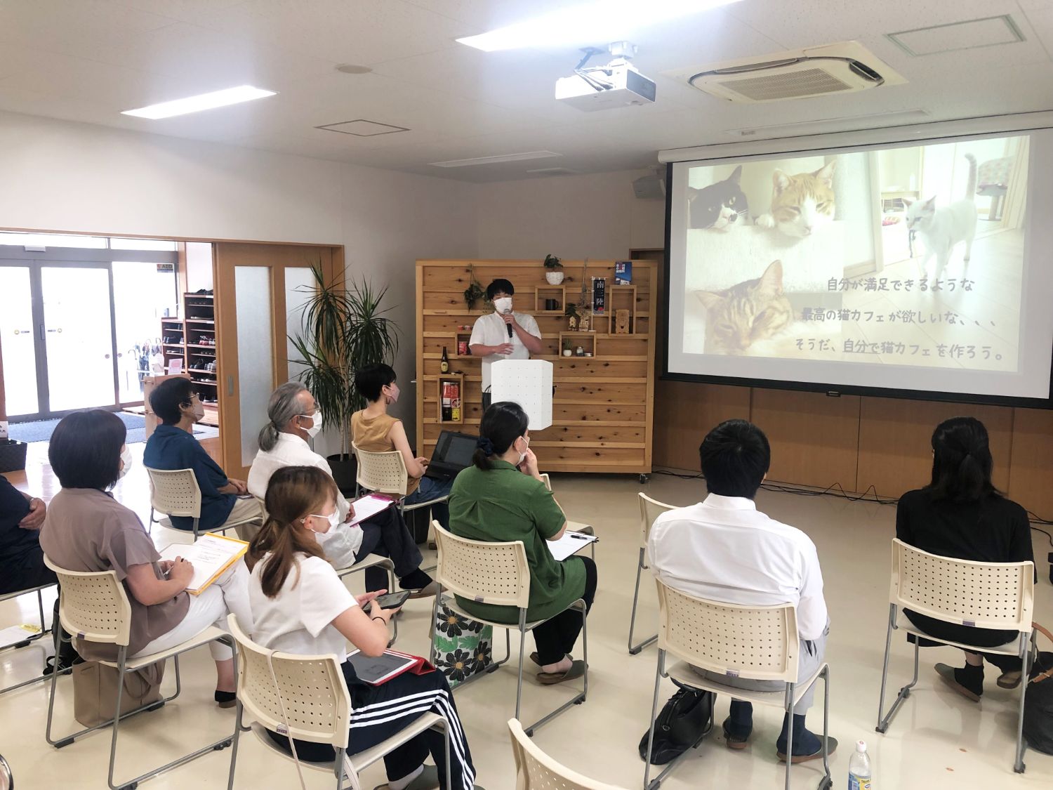 白いシャツを着た男性が前方の演台でマイクを持ってプレゼンを行い、スライドには猫カフェの構想が映し出されており、複数の参加者が椅子に座って真剣に発表を聞いている様子の写真