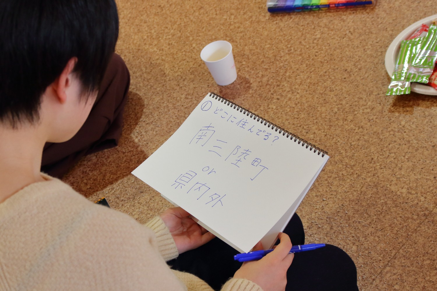 青ペンで「どこに住んでる？ 南三陸町 or 県内外」と書かれたスケッチブックを床に座って見ている若い女性を後方から撮影した写真