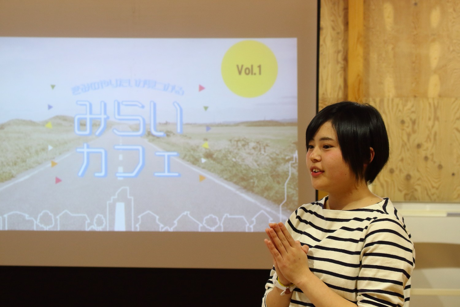 木の壁と「みらいカフェ Vol.1」と書かれたロゴが映っているプロジェクターのスクリーンの前で、ボーダーの服を着た女性が両手を合わせながら話している写真