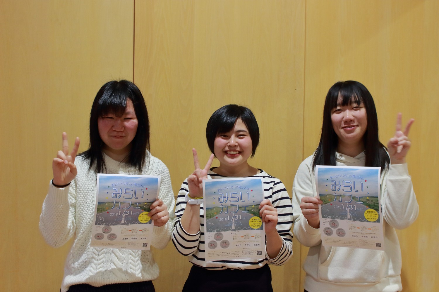 壁の前に並んだ3人の女性が「みらいカフェ」と書かれたチラシを持ってピースサインをしている写真