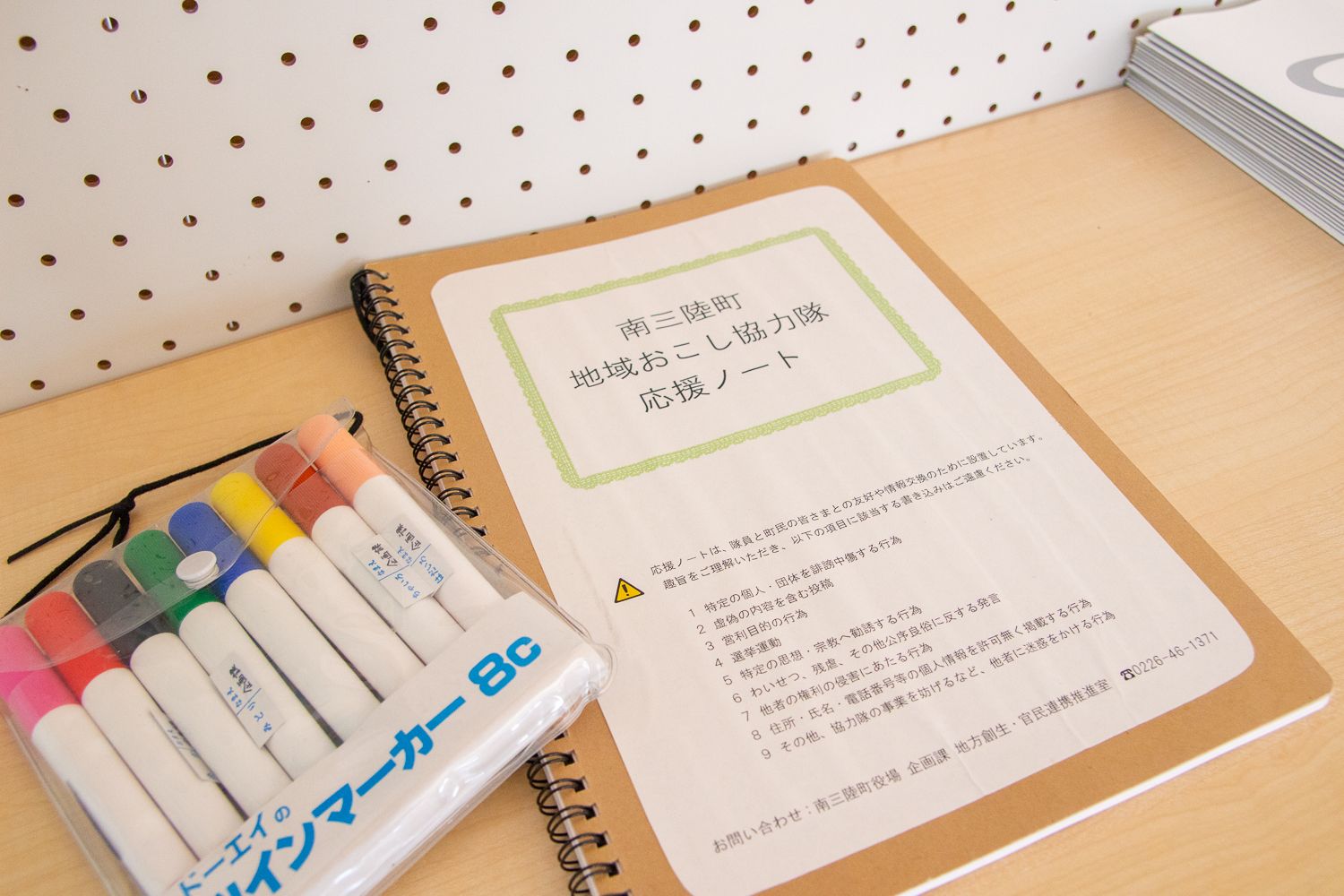 「南三陸町 地域おこし協力隊 応援ノート」と書かれたノートとカラフルなマーカーが机の上に並べて置かれている写真