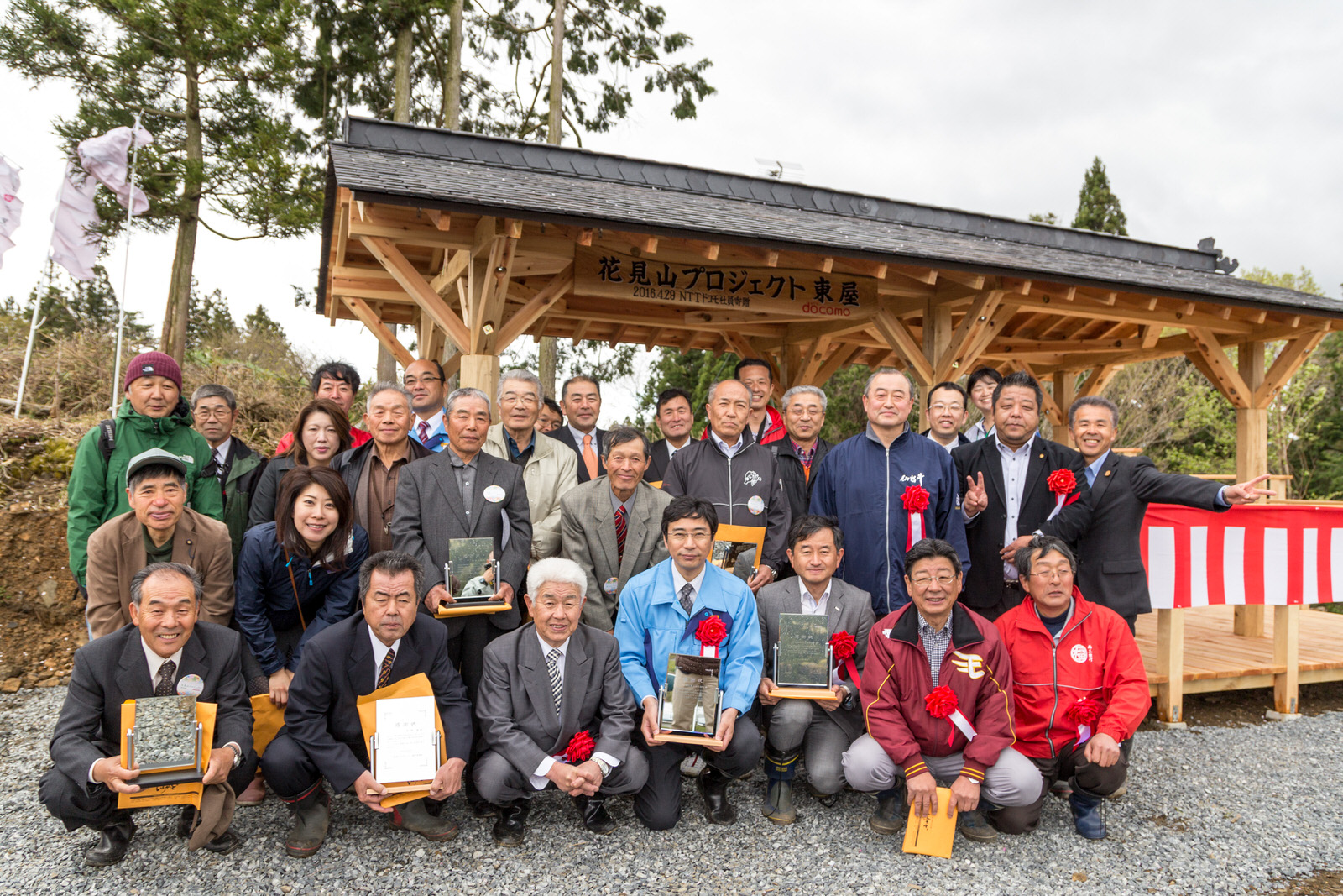 「花見山プロジェクト東屋」と書かれた木の看板が設置され、紅白幕で飾られた東屋の前で大勢の関係者が集まって写した集合写真