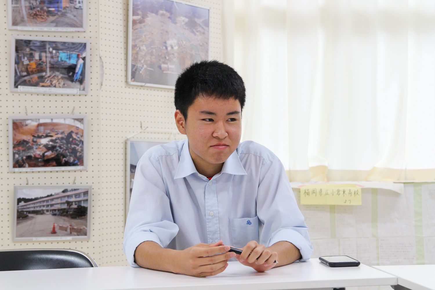 震災の写真がパネルに貼られている室内の長机に座っている木下巧大さんの写真