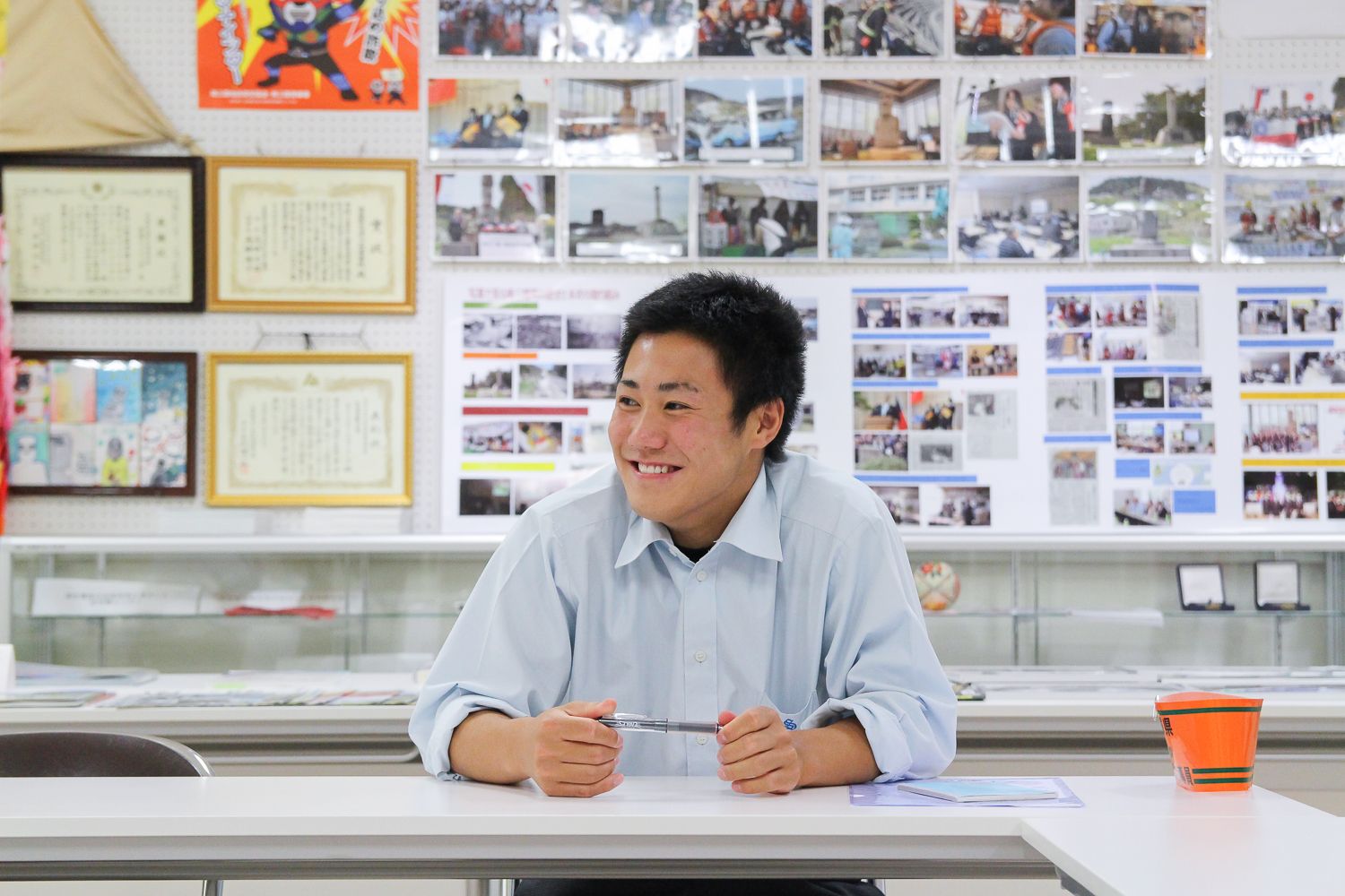 賞状や様々な写真が掲示されている室内の長机に、ペンを手に持ち座っている及川拓海さんの写真