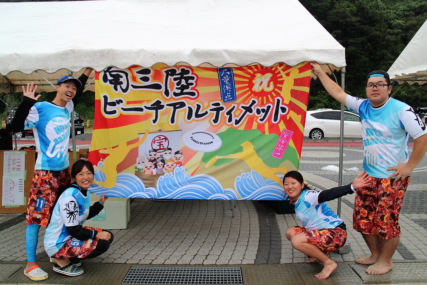 「南三陸ビーチアスティメット」の横断幕の前で実行委員のみなさん4人が笑顔でポーズをしている写真